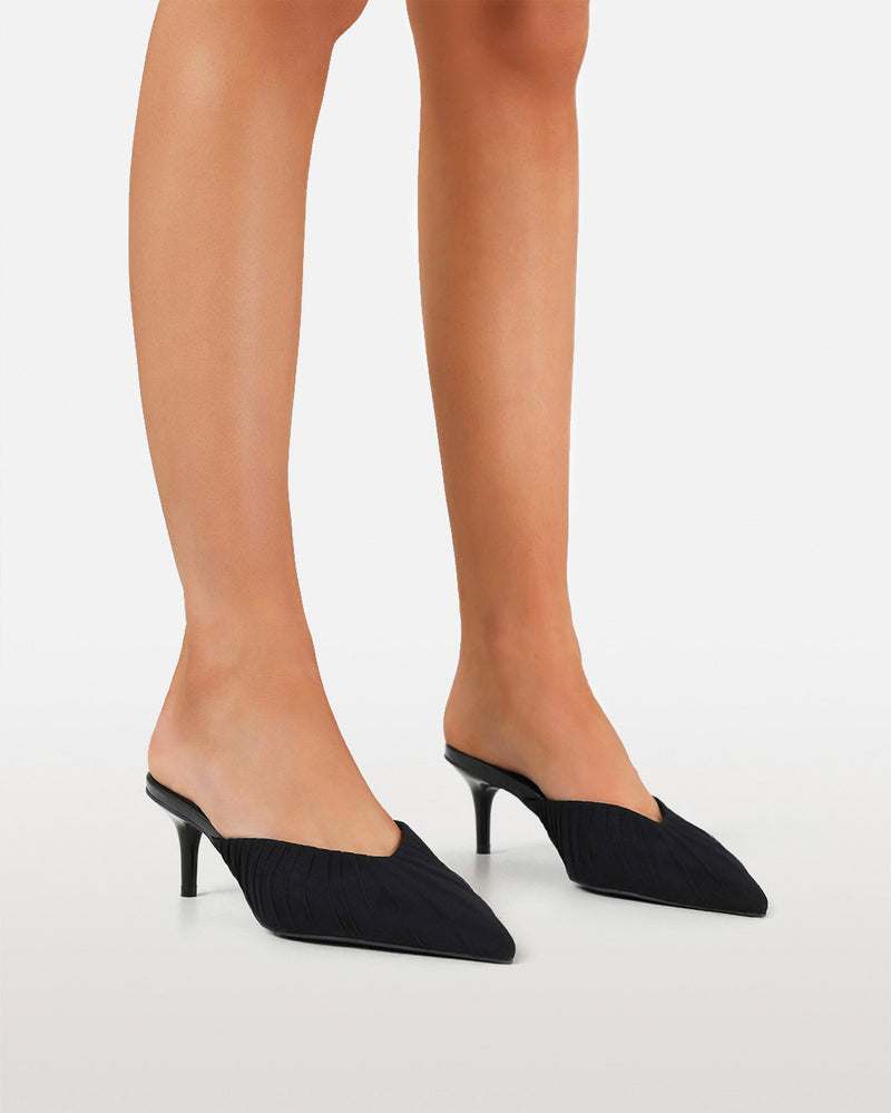 Black pointed-toe mule heels with a kitten heel, shown on a model's feet against a light gray studio background.